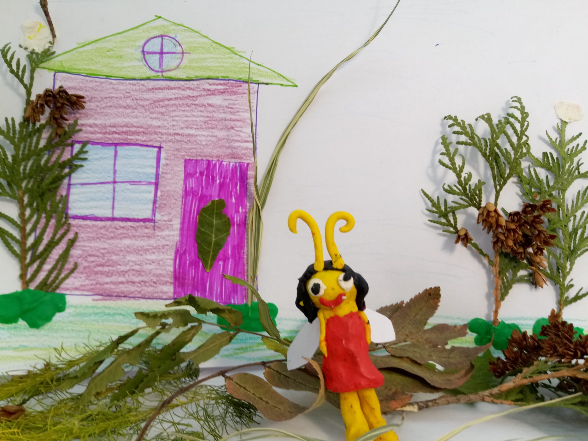 На фото изображена стрекоза из жёлтого пластилина в красном платье. Она стоит на фоне розового дома с ярко зеленой крышей. Крылышки у стрекозы сделаны из белой бумаги. Справа дерево из веточки туи. Слева тоже дерево. 