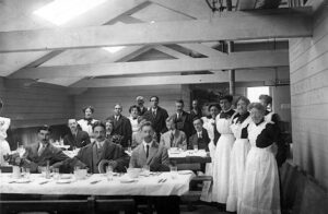 Мужчины в костюмах за обеденными столами и стоящие женщины в черныхдлинных платьях и белых фартуках