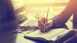 ноутбук, очки и рука, пишущая в тетради